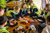 A group of kids in high-vis jackets gather round objects on a table