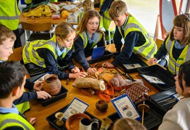 A group of kids in high-vis jackets gather round objects on a table
