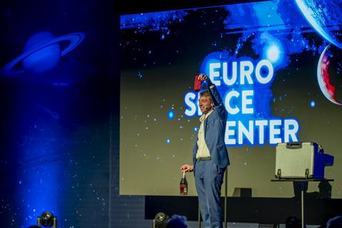 A man on stage holds up a red object at the Euro Space Center